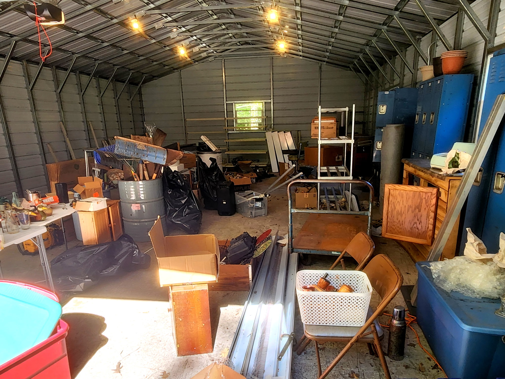Steel outbuilding before the cleanout, filled with boxes, barrels, bags, furniture and household items beside a row of blue lockers.